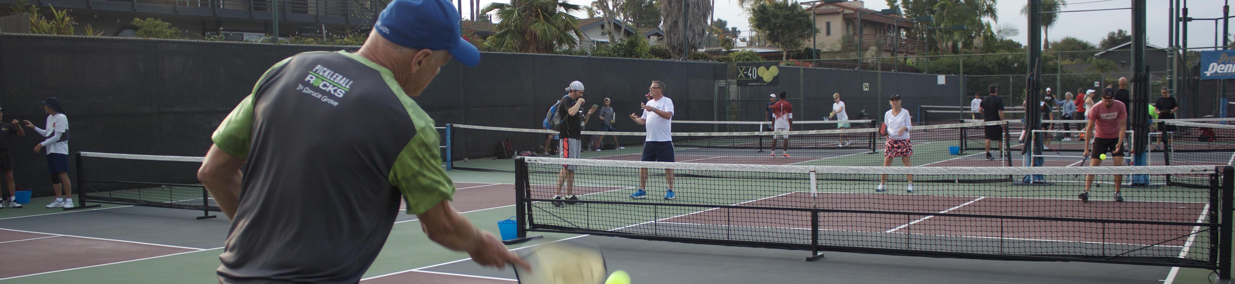 View of pickleball courts. Man wearing gray shirt is returning a serve. Two people are on the other end of the court also warming up. There are some people standing around in the background on various courts either chatting or warming up.